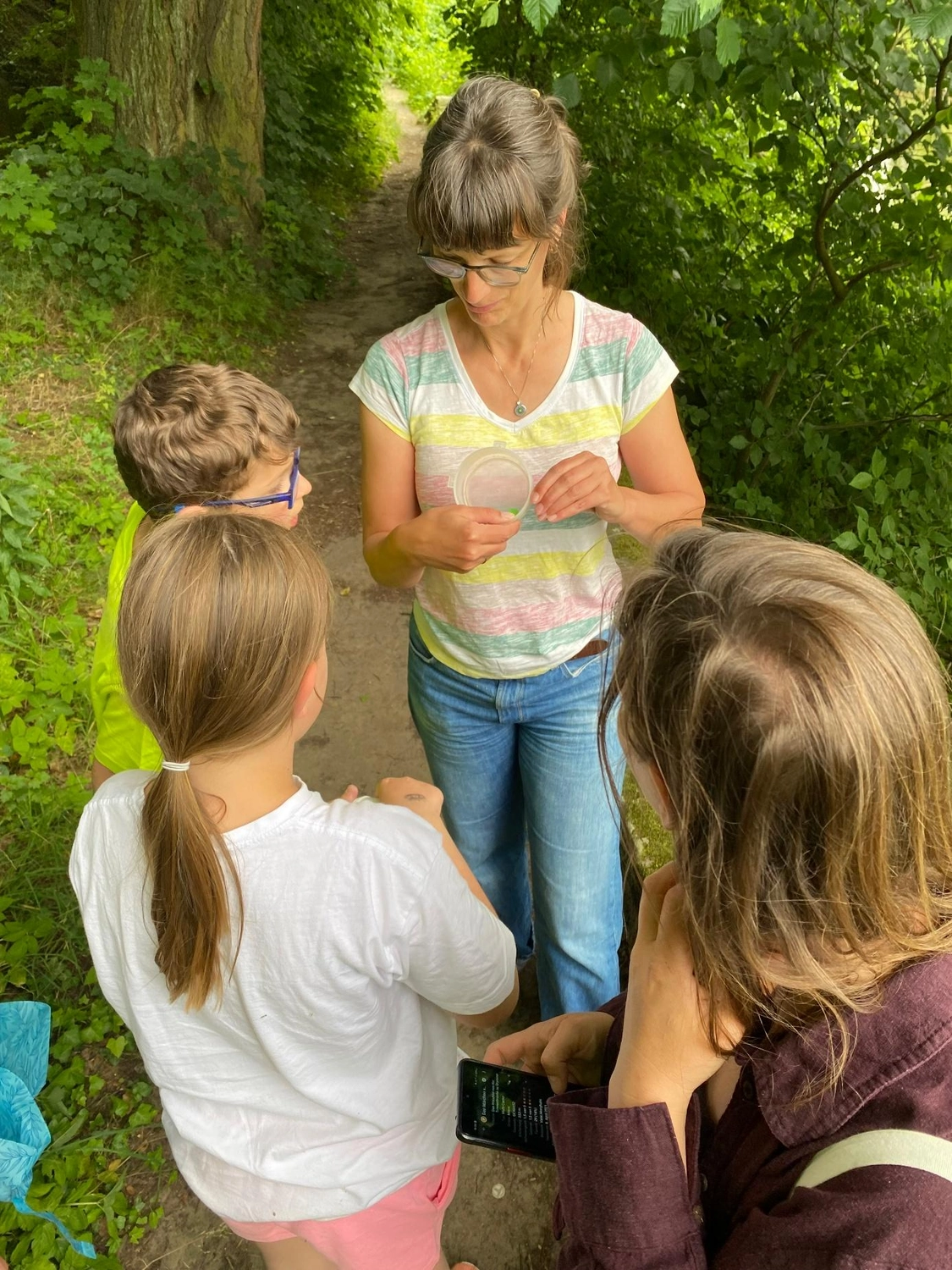 Netzwerk Colditz e.V. - Beziehung. Gestaltung. Entwicklung - Eine Frau mit Brille und gestreiftem Hemd steht auf einem Waldweg und hält drei Kindern einen kleinen, durchsichtigen Behälter hin. Die Kinder, von hinten oder von der Seite zu sehen, beobachten sie neugierig bei einer Lernaktivität im Freien.