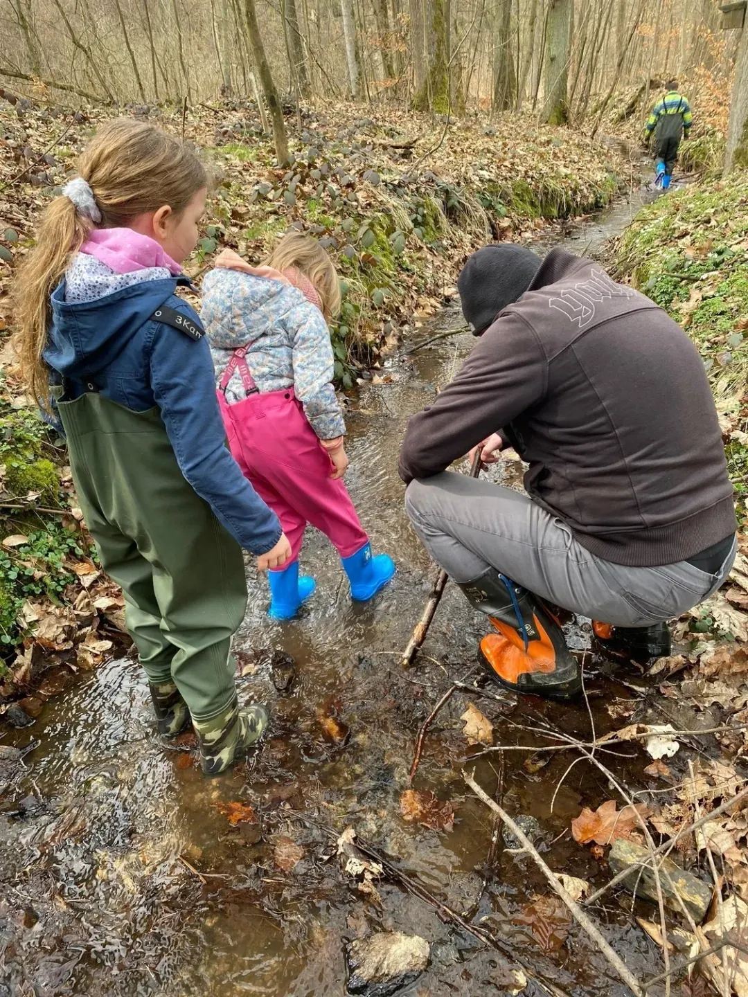 Eine erwachsene Person und zwei Kinder erkunden in Outdoor-Kleidung gemeinsam einen flachen, steinigen Bach im Wald.